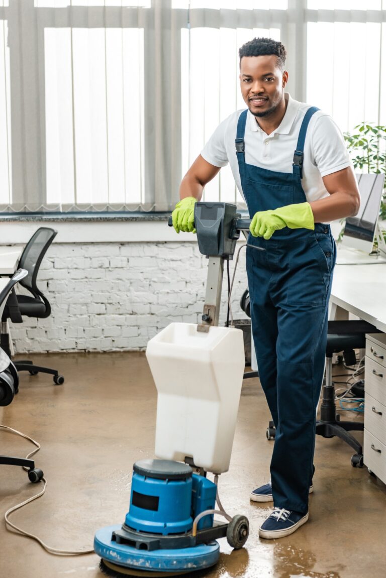 smiling african amercan cleaner looking at camera while washing floor with cleaning machine